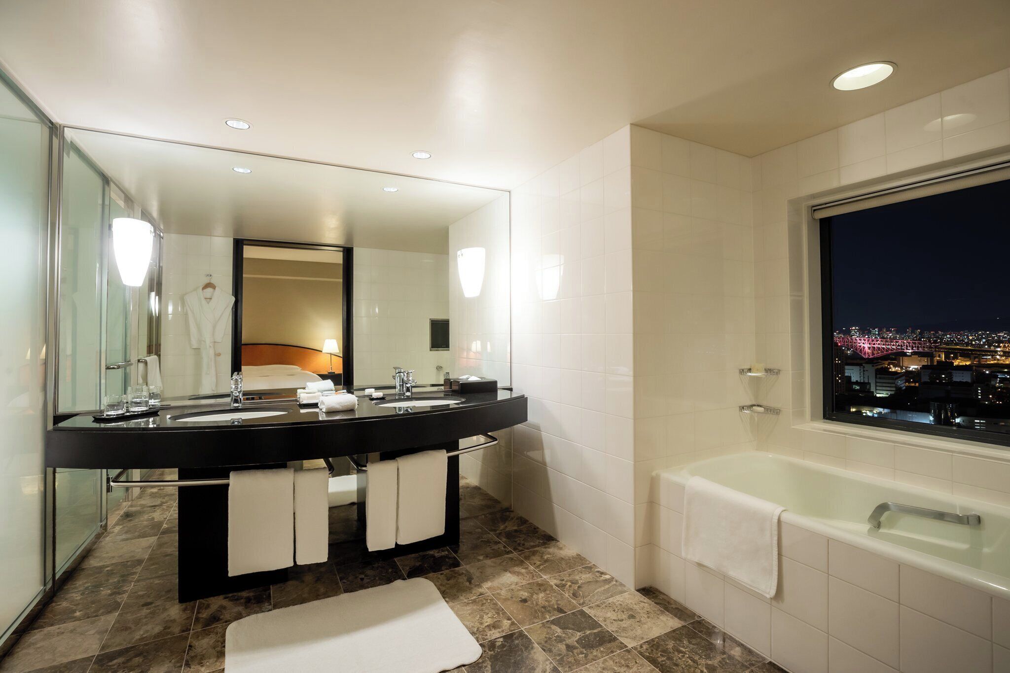 A modern bathroom with a large mirror, dual sinks, a bathtub, and a city view through a window.