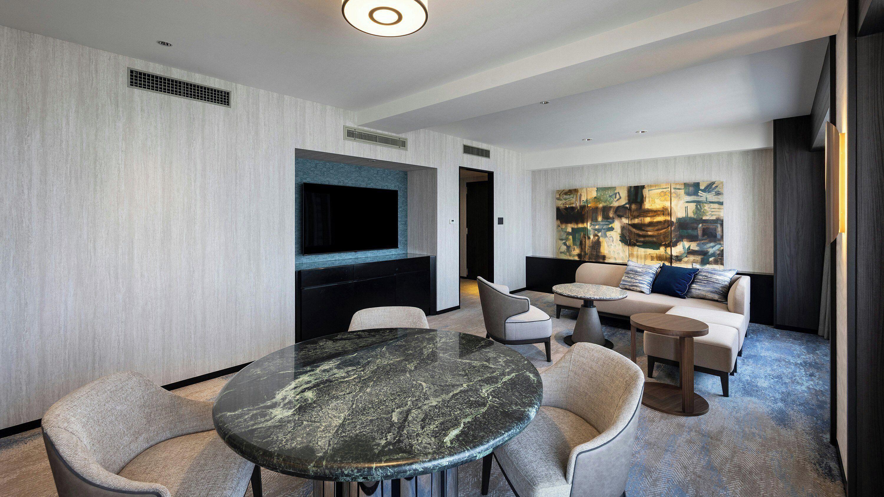 Une chambre d’hôtel moderne avec une table à manger en marbre, des sièges confortables, un téléviseur à écran plat et un grand tableau abstrait accroché au mur.