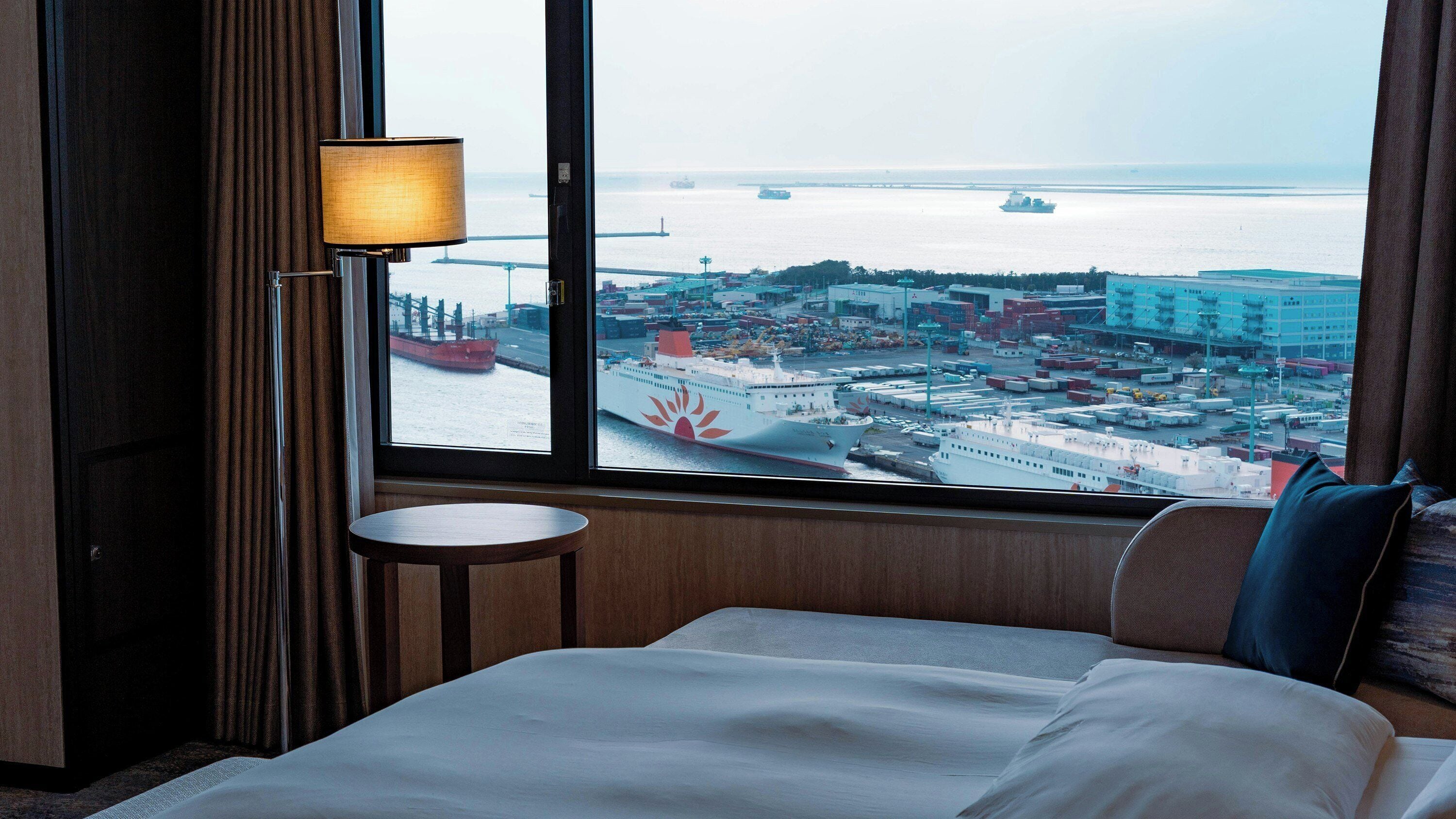 A hotel room with a large window offering a view of a port with ships and industrial buildings.