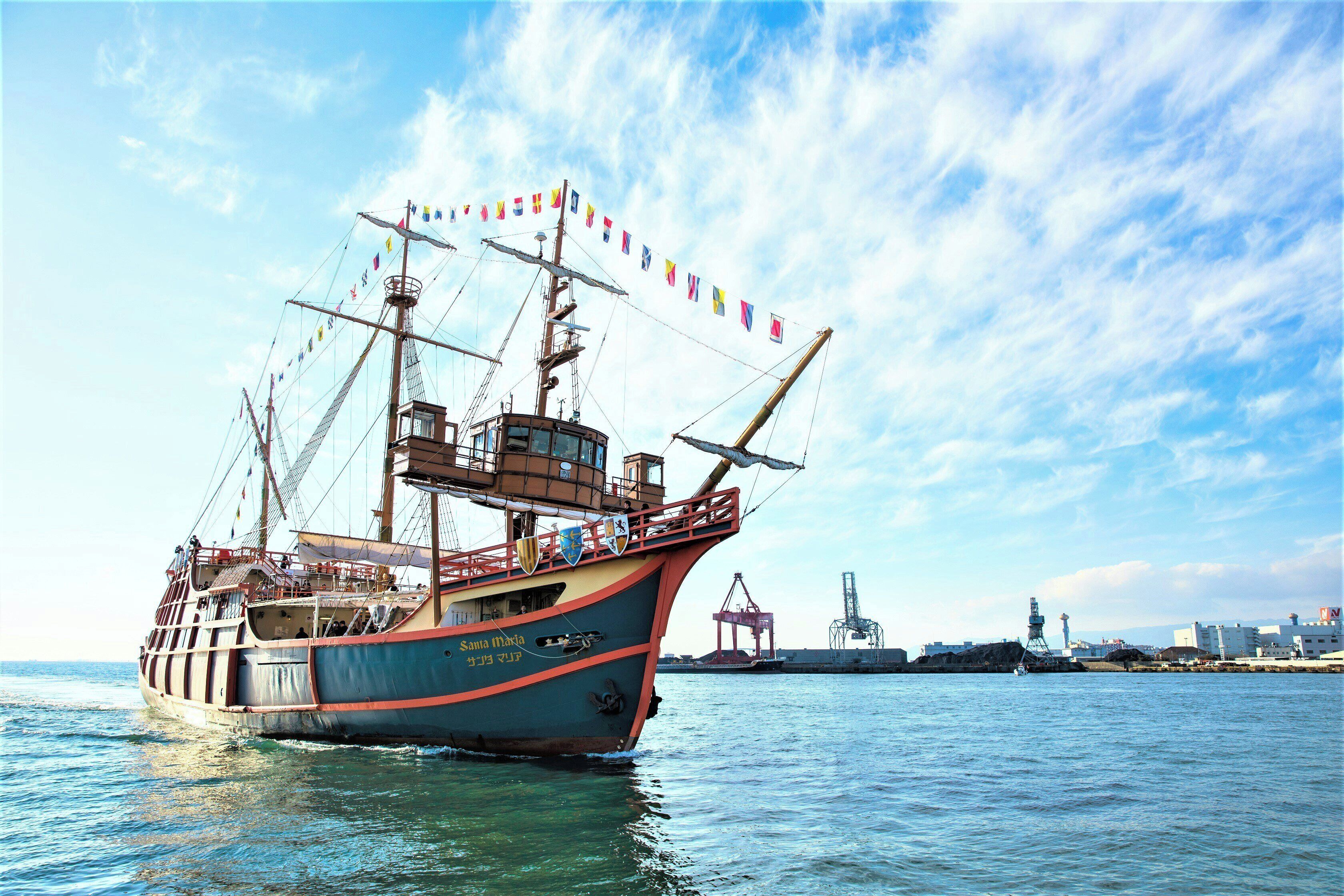 A large ship with multiple decks and a cabin structure, docked at a port with industrial buildings and cranes in the background.