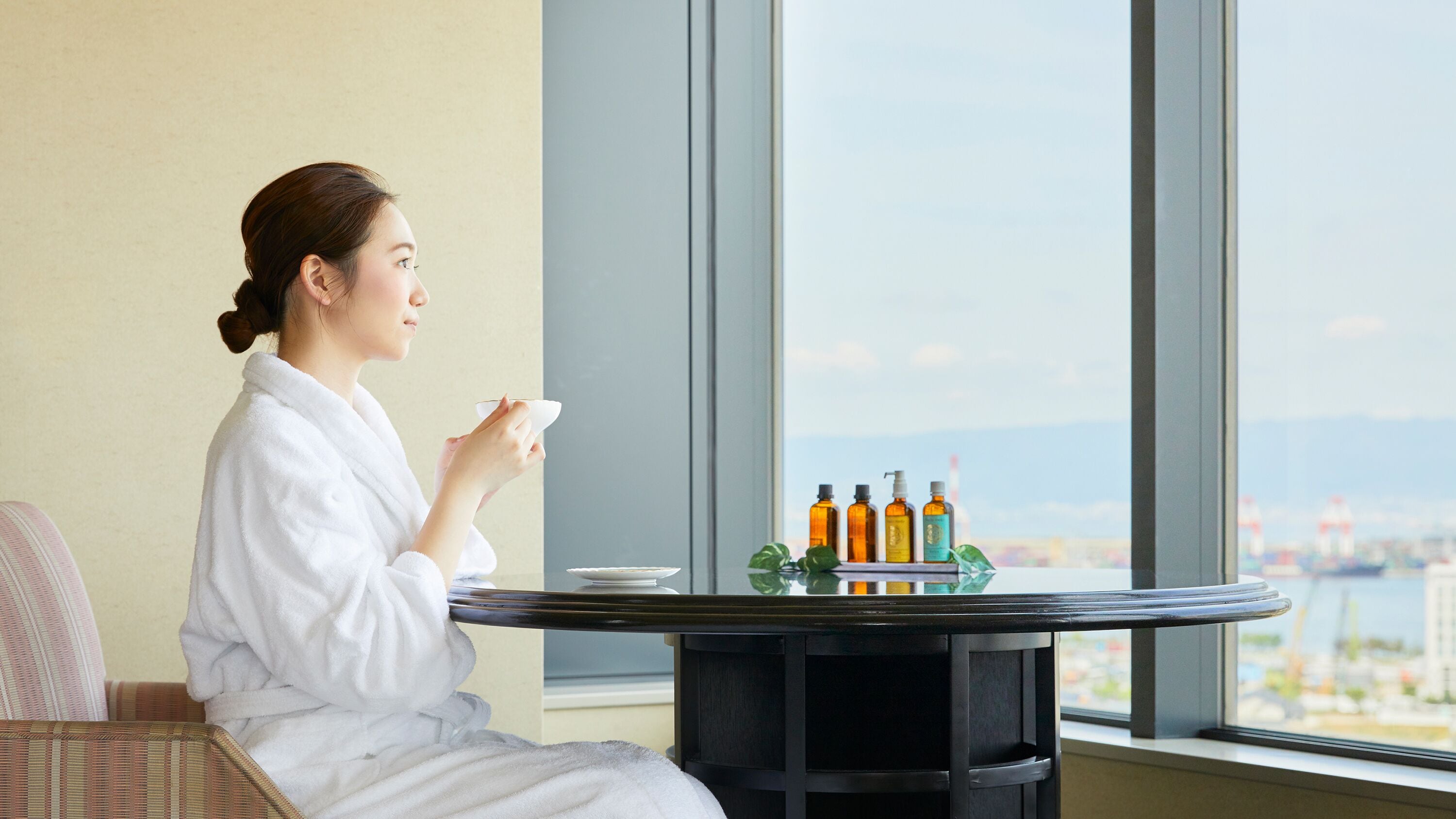 Una persona con bata blanca sentada junto a una ventana con vistas a un paisaje urbano, una mesa con botellas y un plato.