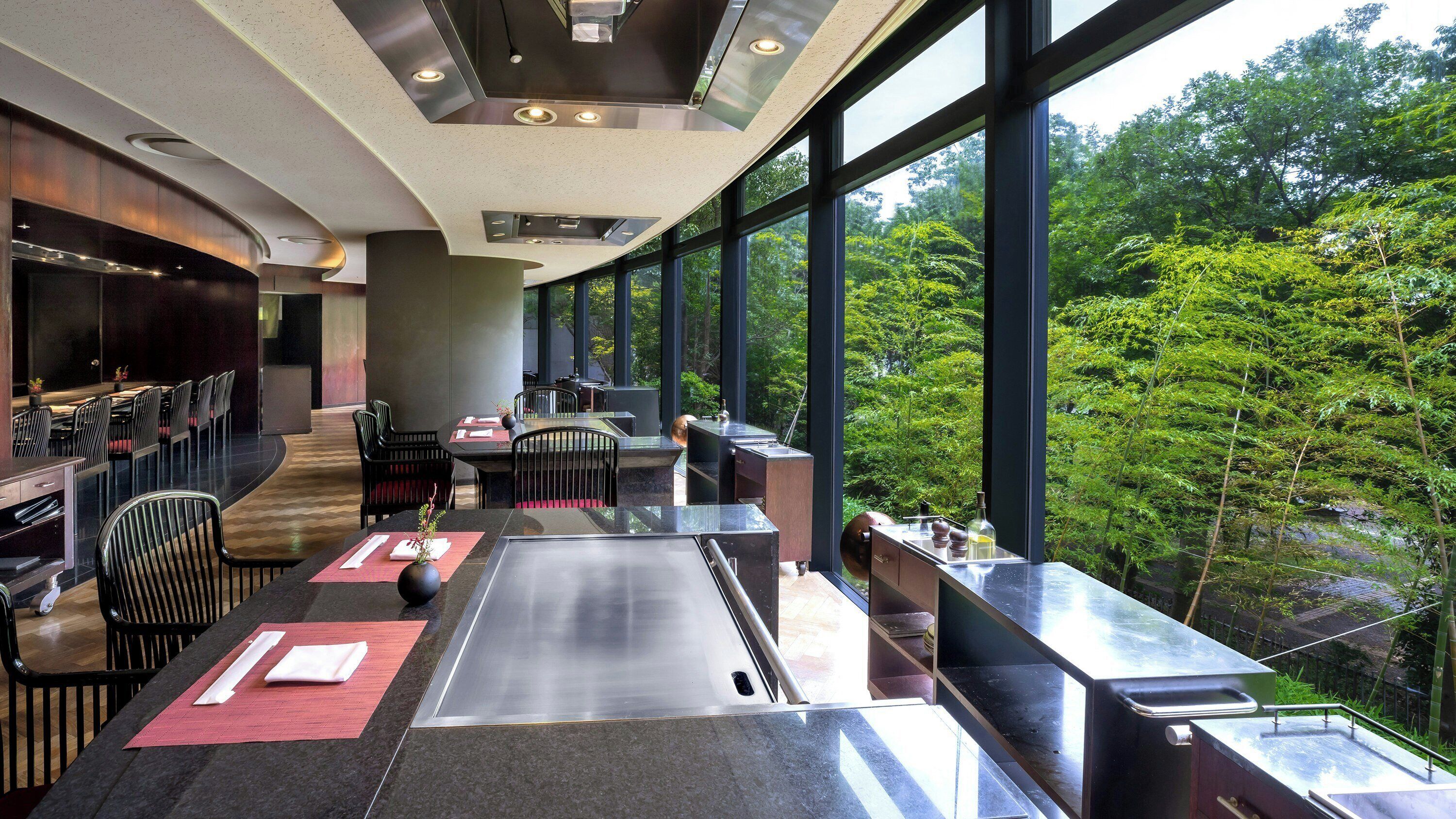 A modern restaurant with a long counter, black chairs, and a view of lush greenery outside.