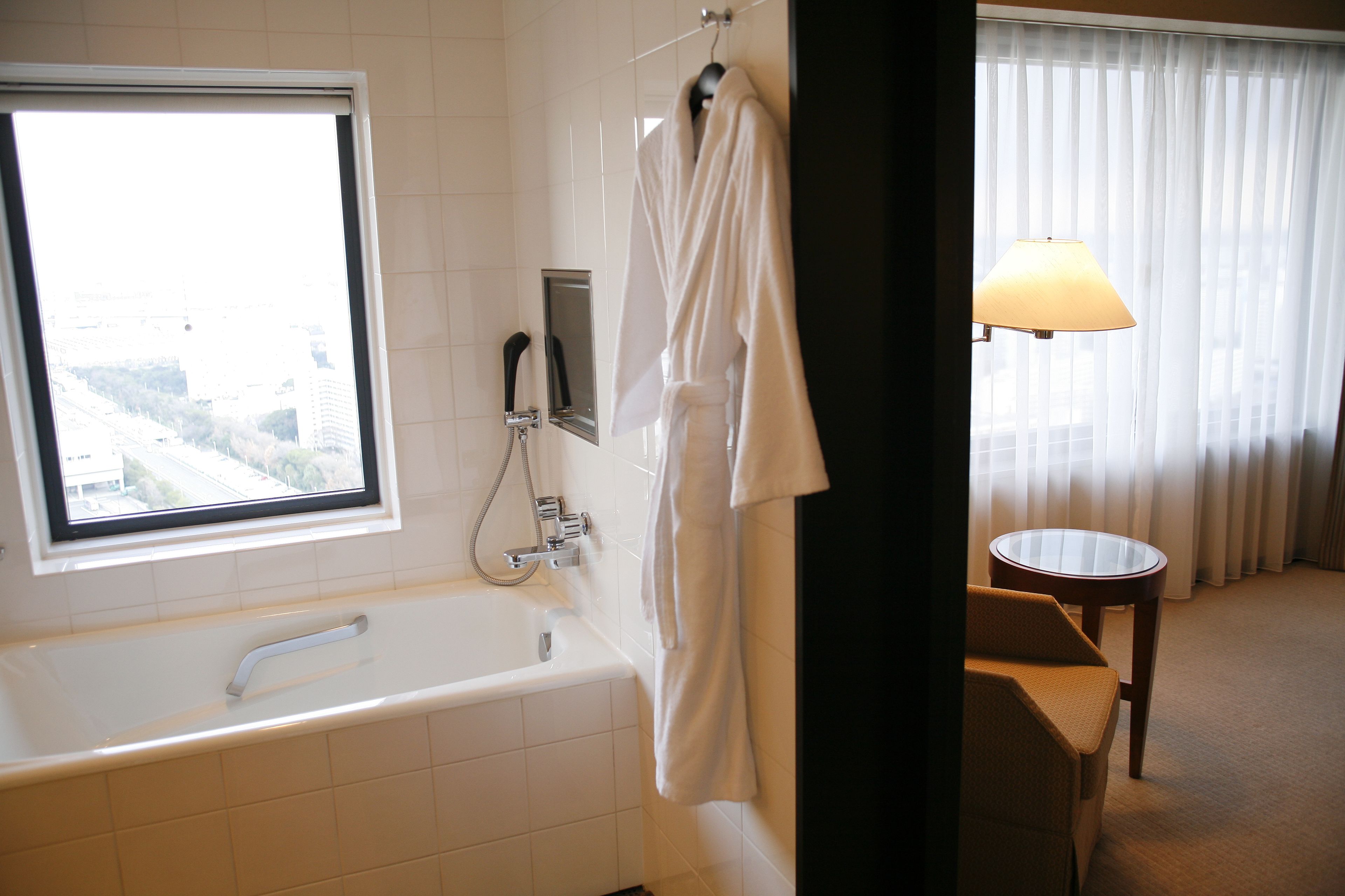 A bathroom with a bathtub, a mirror, a robe, a window, and a lamp.