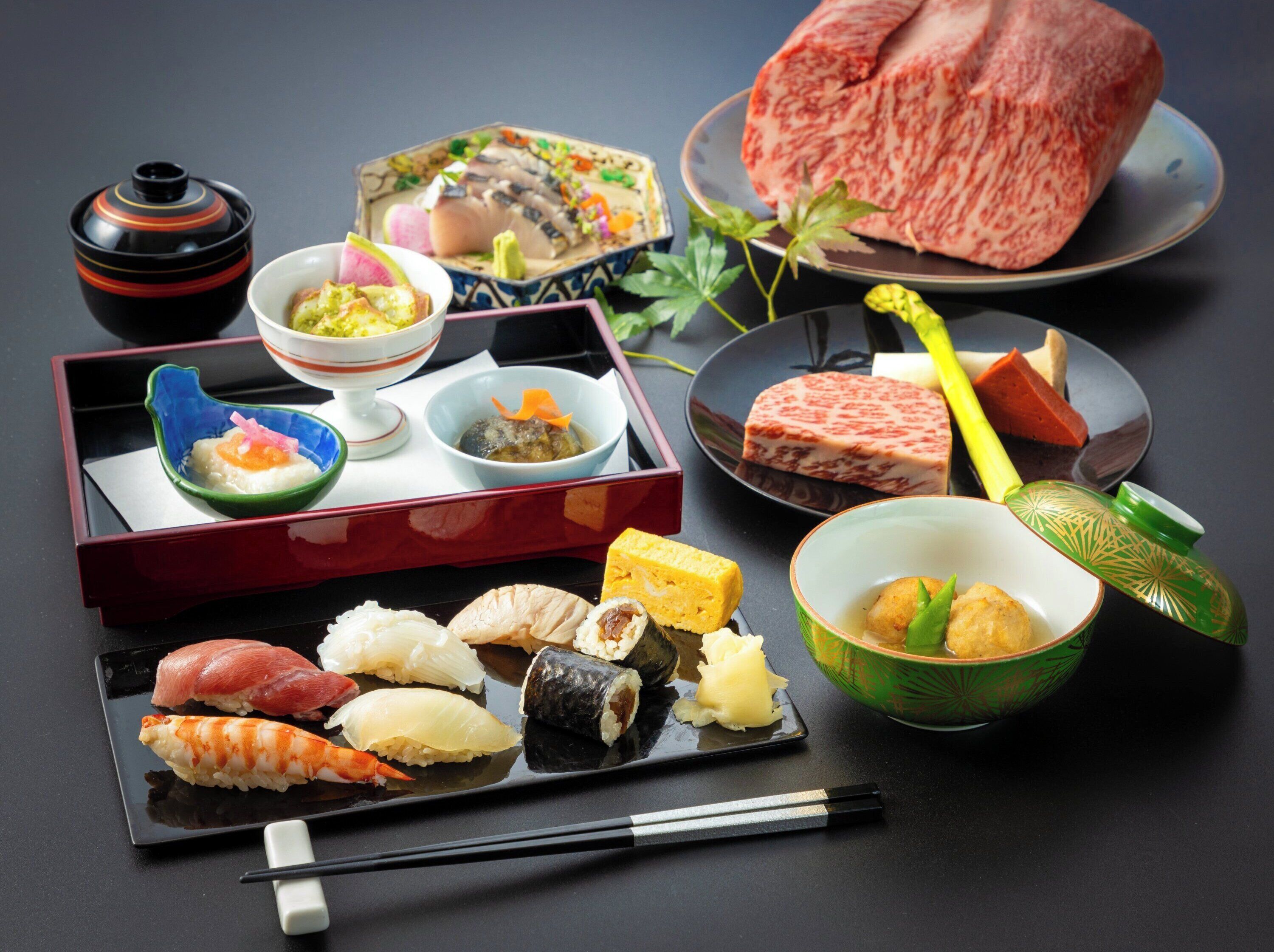 A variety of Japanese dishes including sushi, sashimi, and a large piece of raw beef, arranged on a dark table.