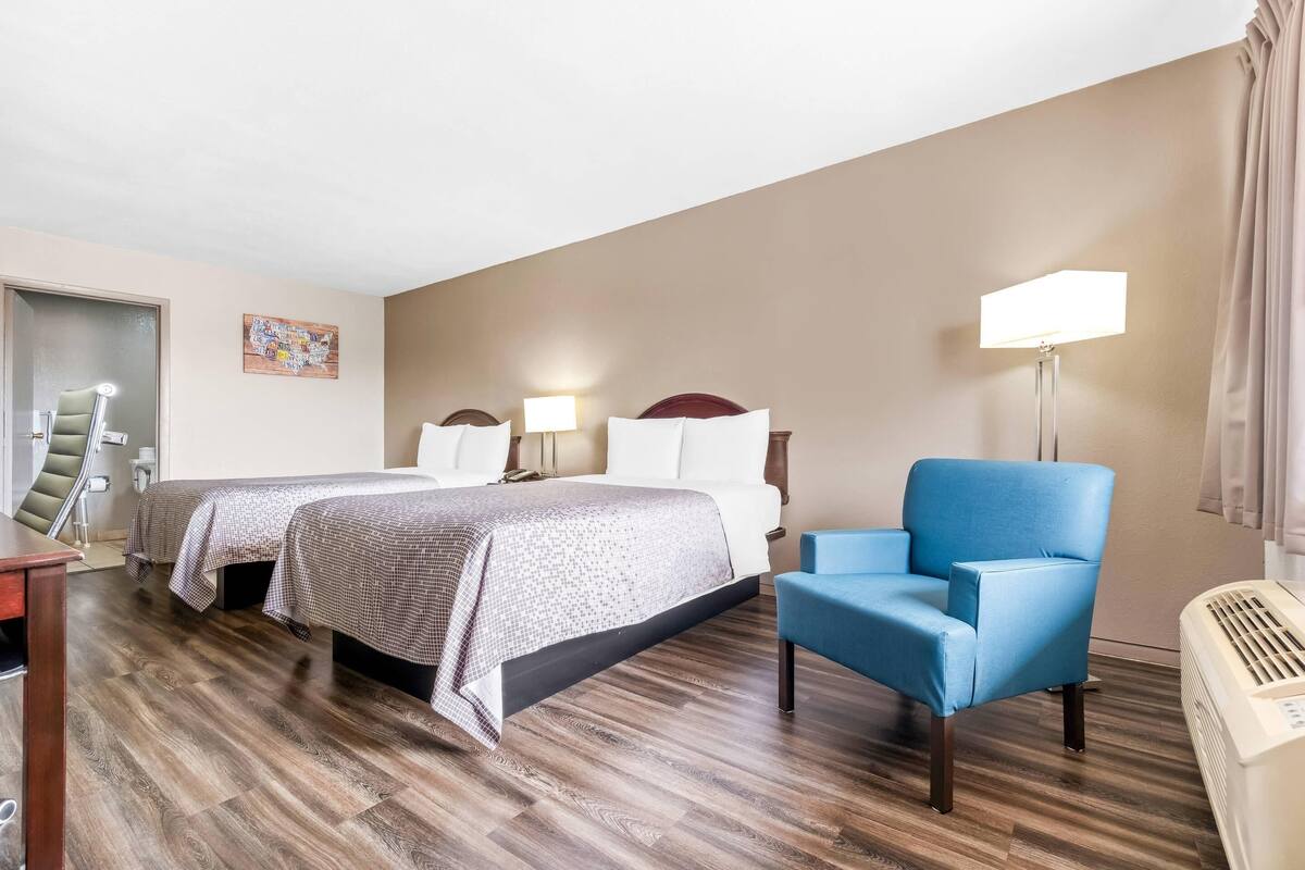 A hotel room with two beds, a blue armchair, a desk, a lamp, and a wall-mounted air conditioner.