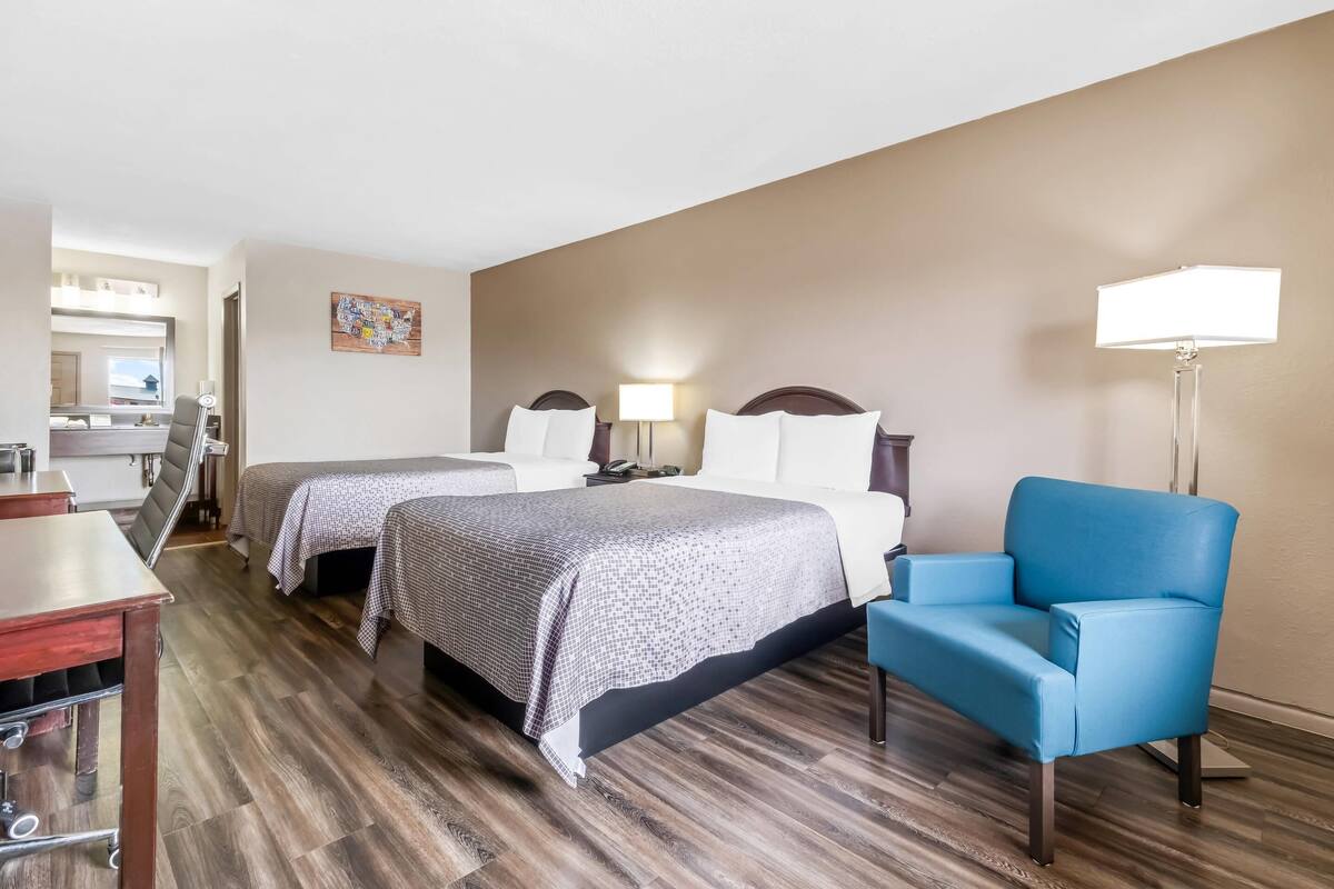A hotel room with two beds, a desk, a chair, and a lamp.
