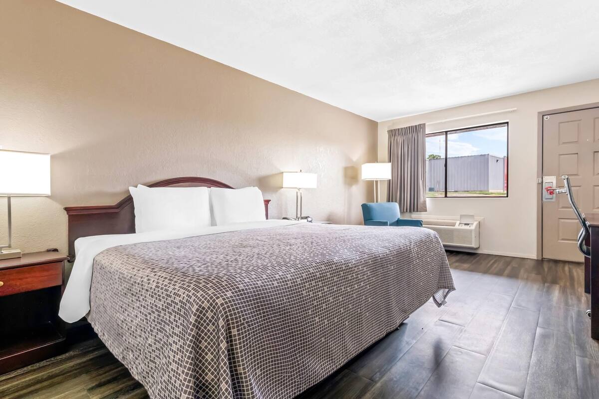 A hotel room with a large bed, two bedside lamps, a chair, a desk with a lamp, and a window with curtains.