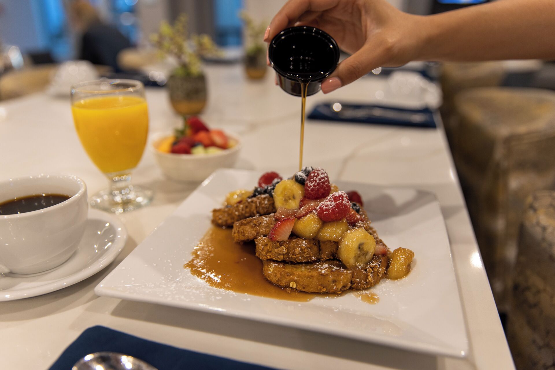 Un plato de desayuno con waffles cubiertos de fruta y jarabe, un vaso de jugo de naranja y una taza de café sobre una mesa de hotel.