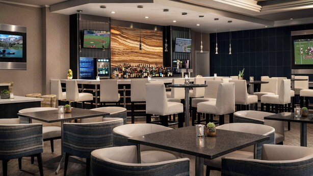 A modern bar area with multiple seating options, a bar counter with a variety of bottles, and two large screens displaying sports games. - Visalia Marriott at the Convention Center (Visalia)