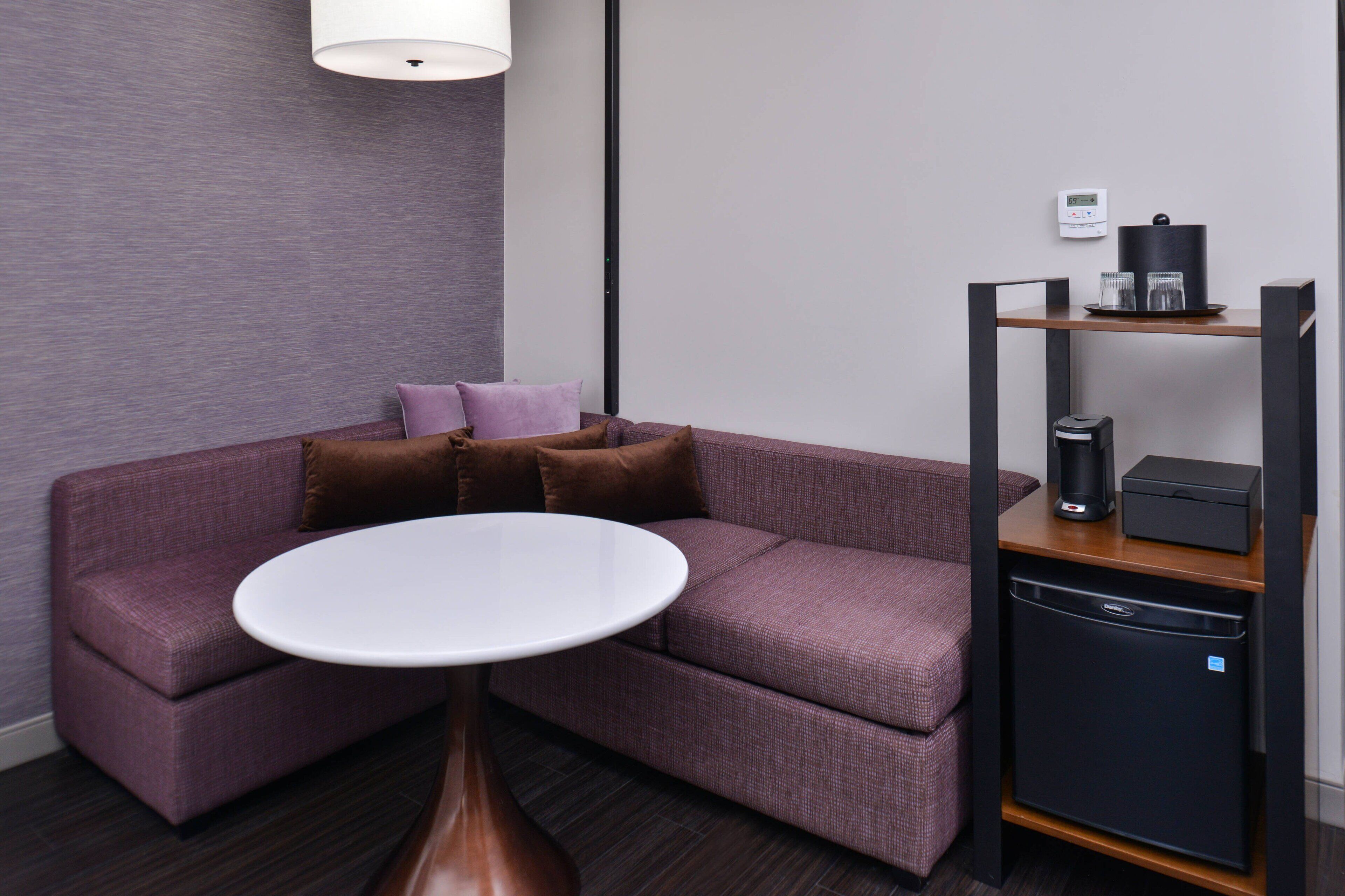 A modern hotel room with a purple sofa, a white round table, a coffee machine, and a mini-fridge.