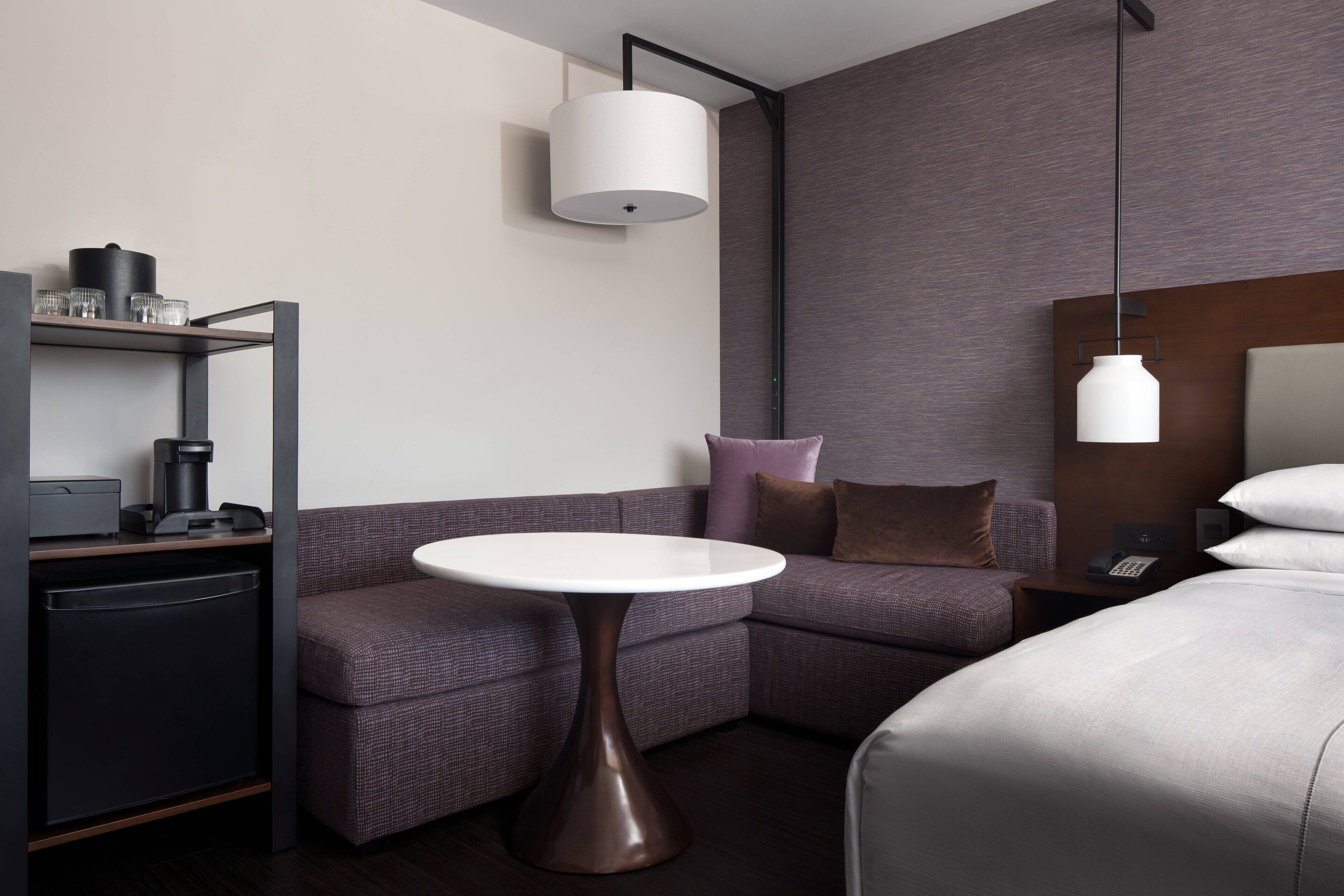 A modern hotel room with a sofa, a small round table, a bed, a nightstand with a phone, and a wall-mounted lamp.