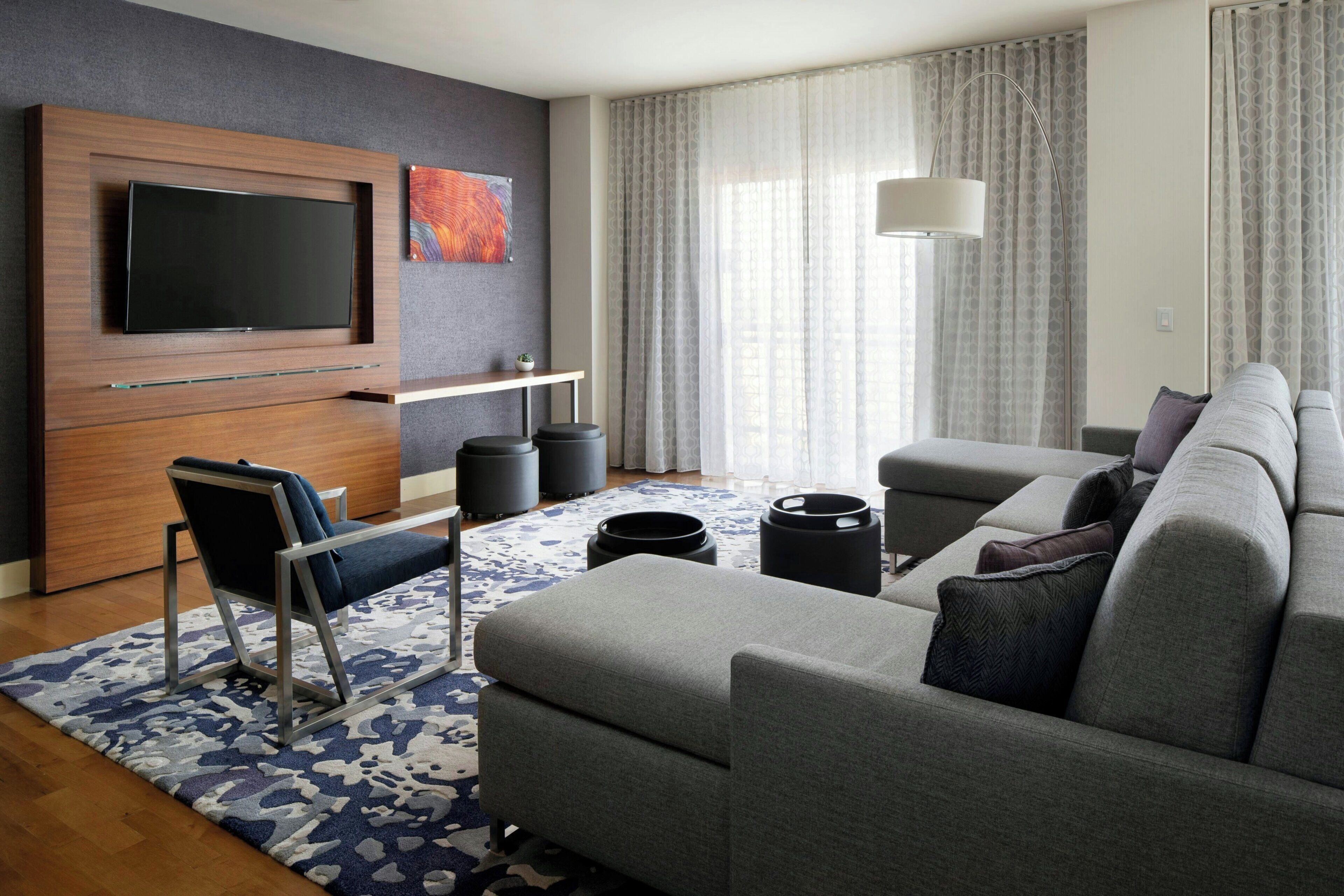 Un salon moderne doté d’un téléviseur à écran plat, d’un canapé gris, d’un tapis à motifs bleus et d’une grande fenêtre avec des rideaux.
