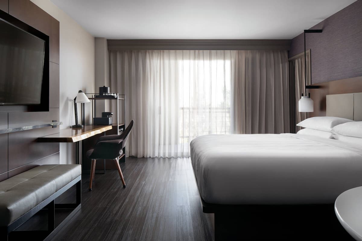A modern hotel room with a large bed, a desk with a chair, a TV, and a window with curtains.