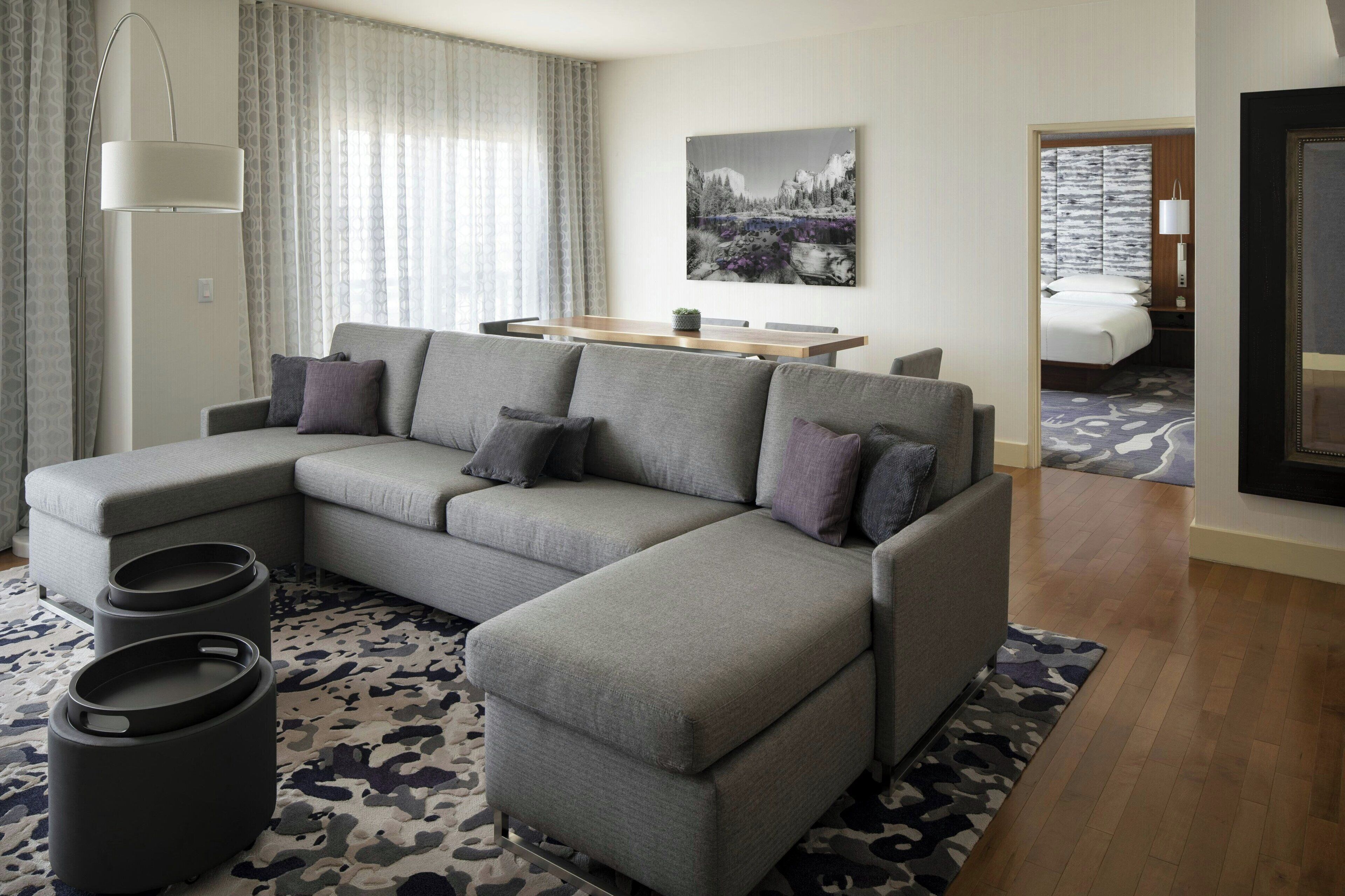 A modern living room with a sectional sofa, a dining table, and a bedroom visible through an open door.