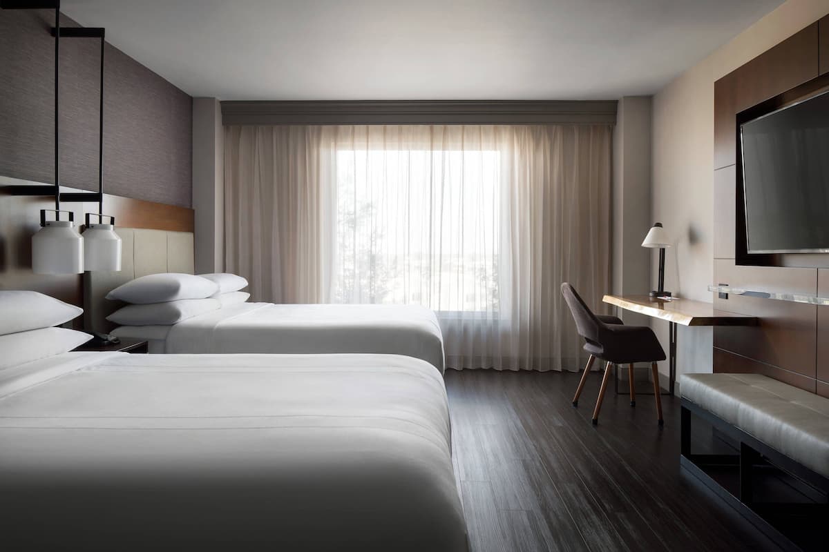 A hotel room with two beds, a desk, a chair, a TV, and a large window with curtains.