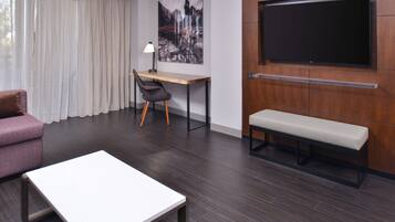 A modern hotel room with a wooden desk, a bench, a sofa, and a large TV mounted on the wall.