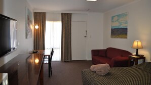 Ein Hotelzimmer mit einem Holztisch, einem roten Sofa, einem bettenbezogenen Bett, einem Fernseher und einem Gemälde an der Wand.