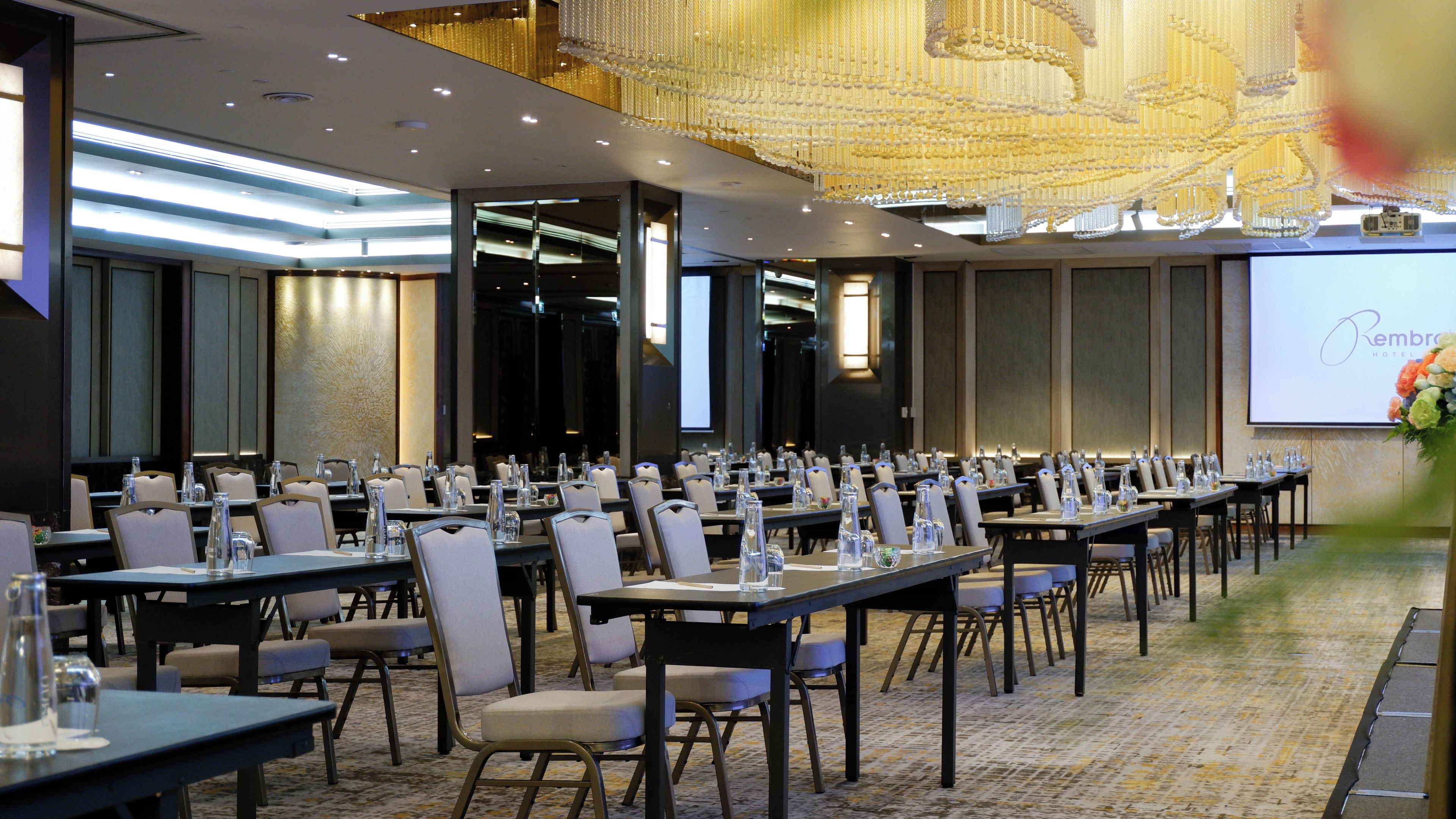 A conference room with rows of tables and chairs, a large screen, and a chandelier.