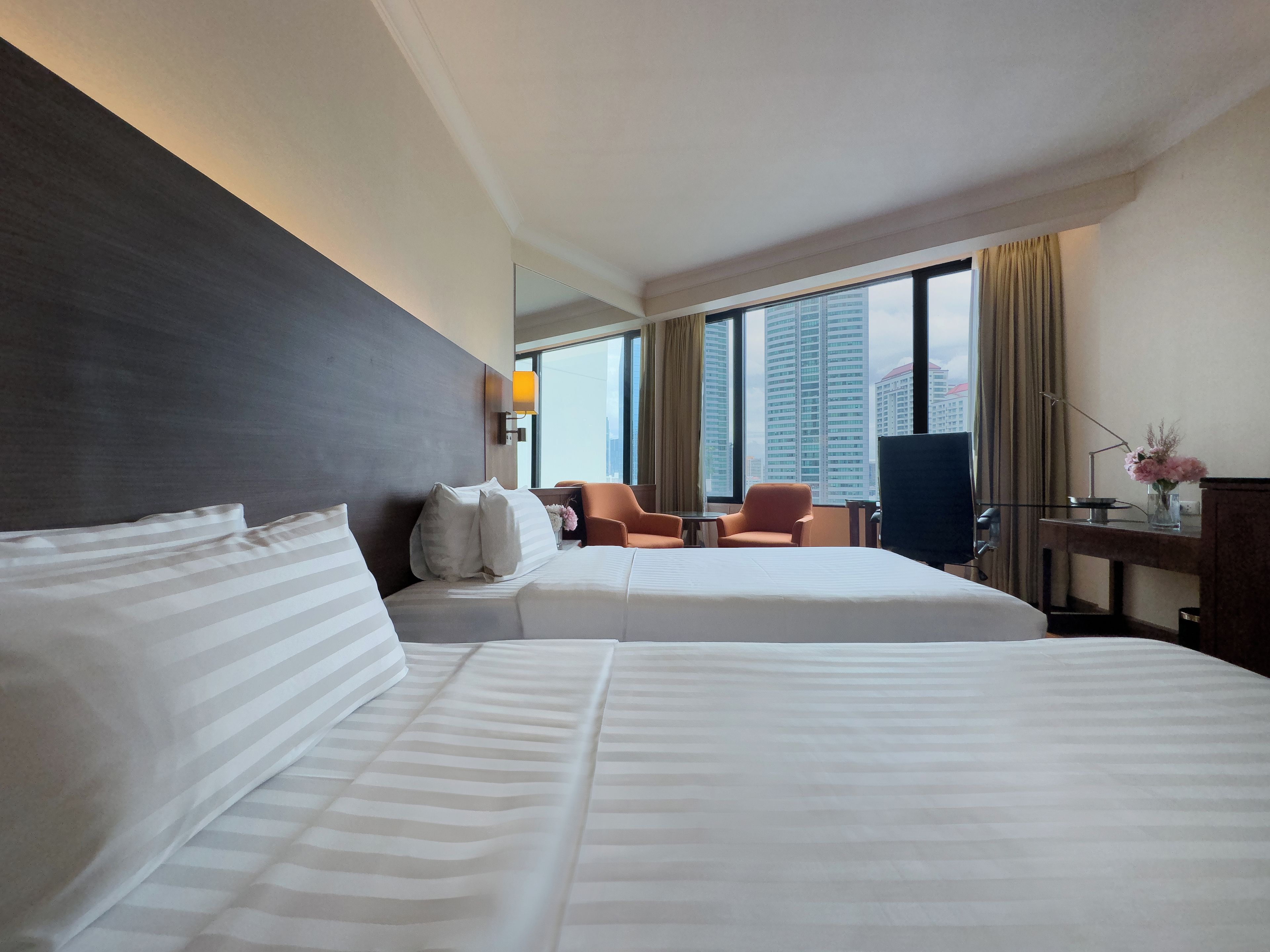 Quarto de hotel com duas camas, uma escrivaninha, uma cadeira e uma janela grande com vista para a cidade.