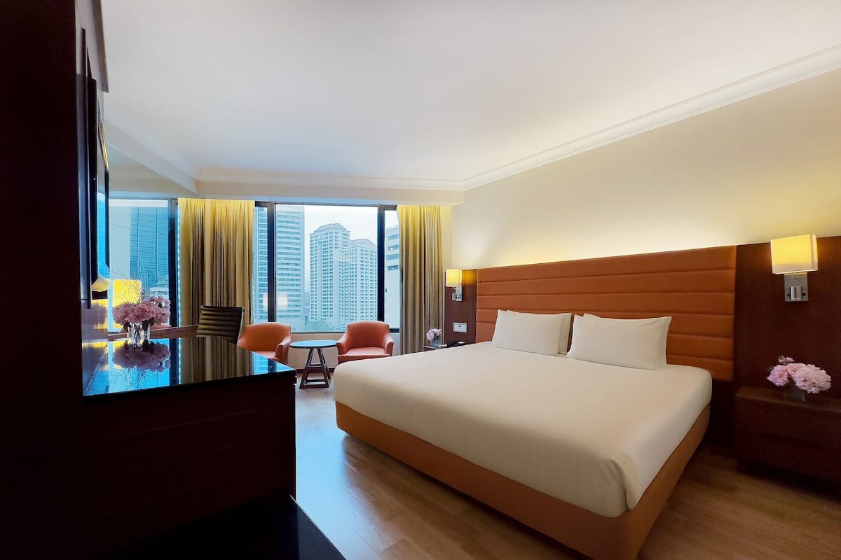 A hotel room with a large bed, a desk with a chair, a small table, and a view of tall buildings through large windows.