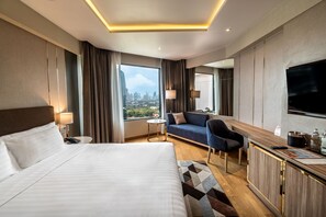 A hotel room with a large bed, a sofa, a desk with a TV, and a city view. - Rembrandt Hotel Bangkok (Bangkok)