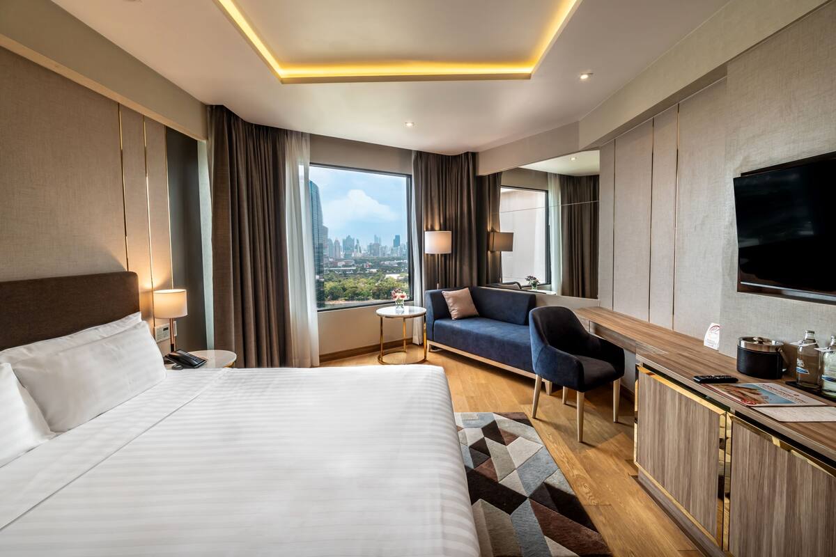 A hotel room with a large bed, a sofa, a desk with a TV, and a city view.