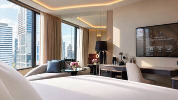 A modern hotel room with a large bed, a desk, a chair, and a TV displaying the hotel's name.