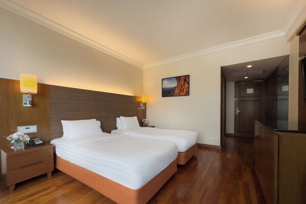 A hotel room with two beds, wooden flooring, a nightstand with a phone, a lamp, and a painting on the wall.
