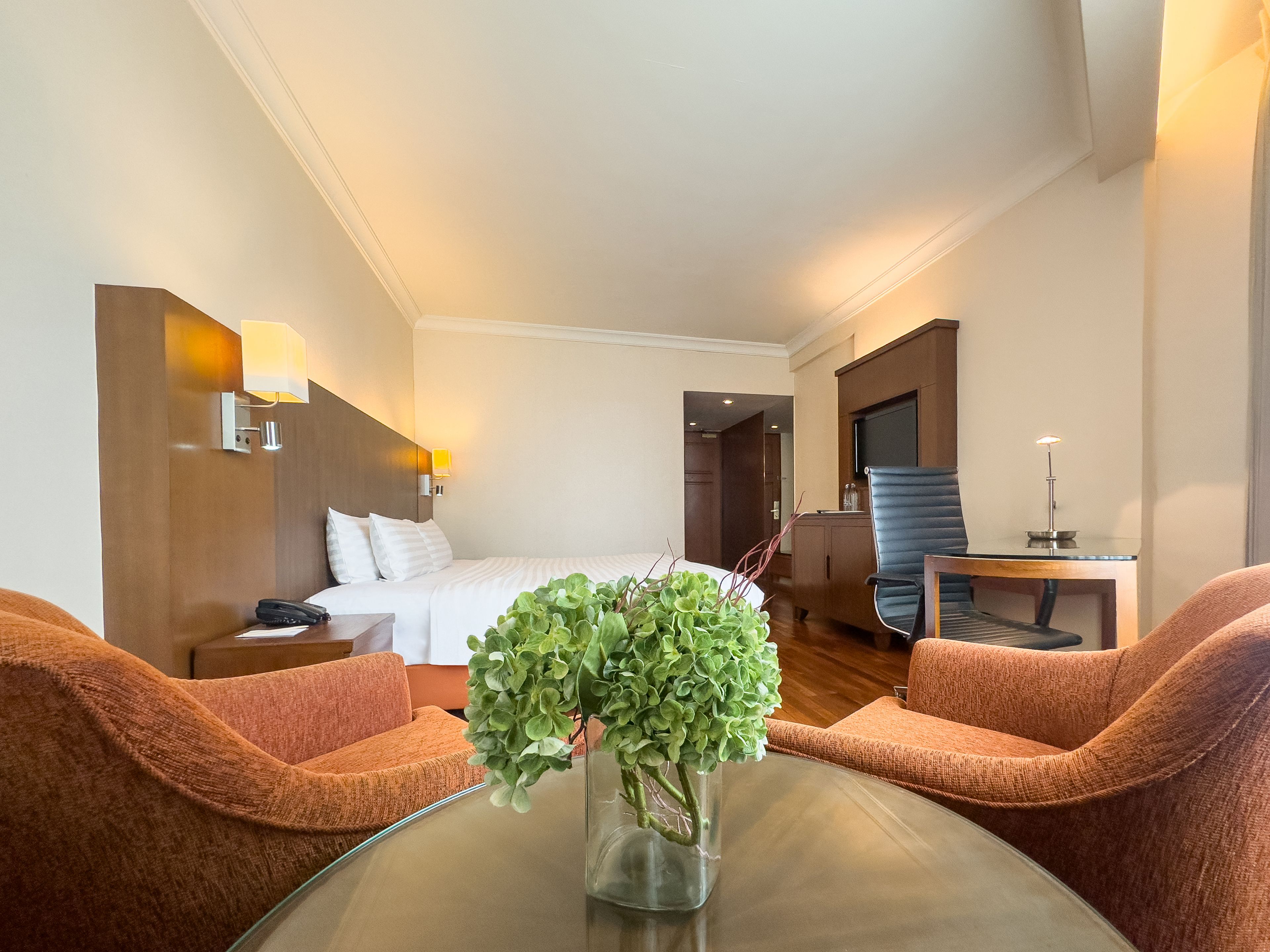 A hotel room with a bed, two armchairs, a glass table with a vase of green plants, a desk with a lamp, and a wooden cabinet.