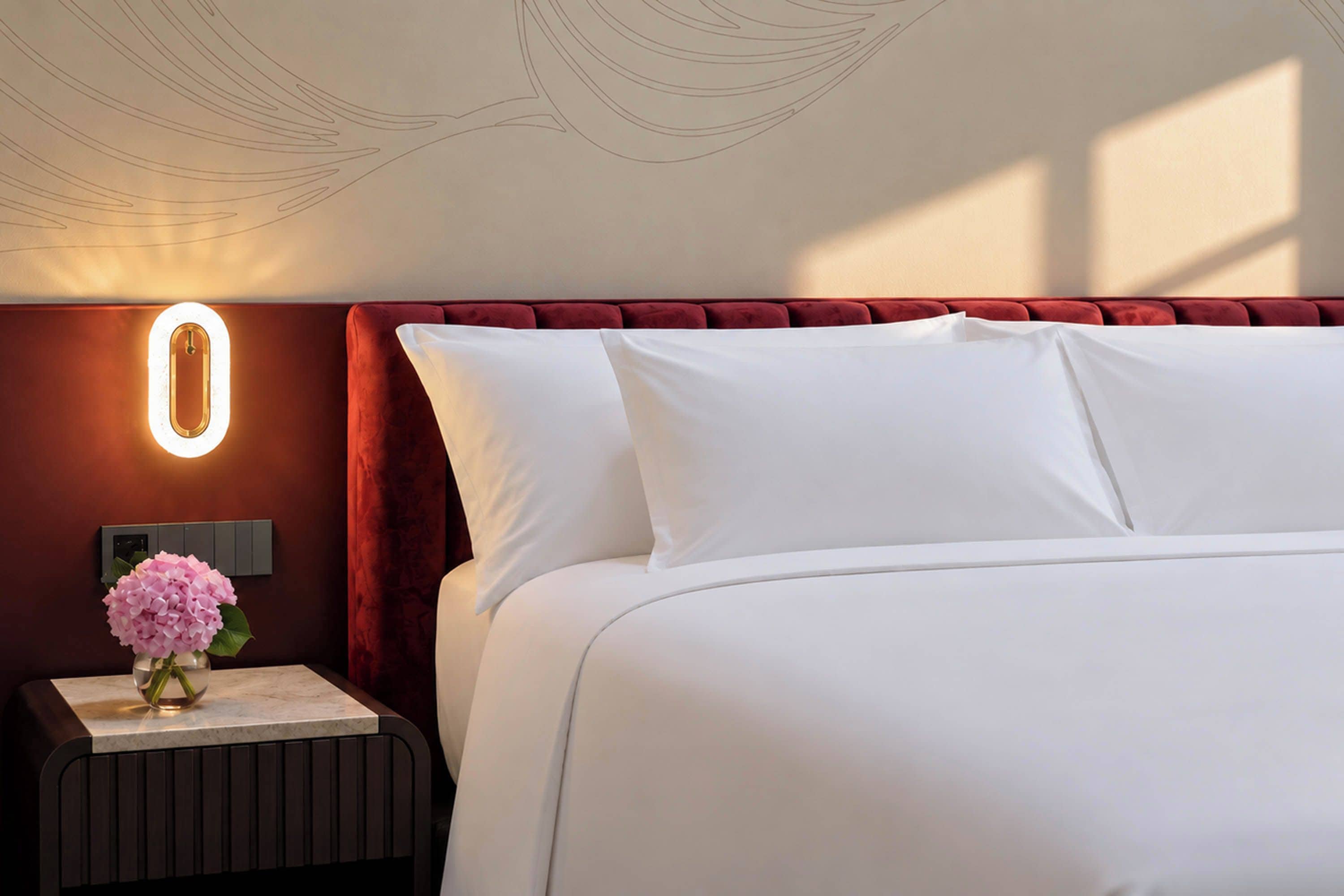 A hotel room with a bed, a nightstand, a vase with flowers, and a wall-mounted light fixture.