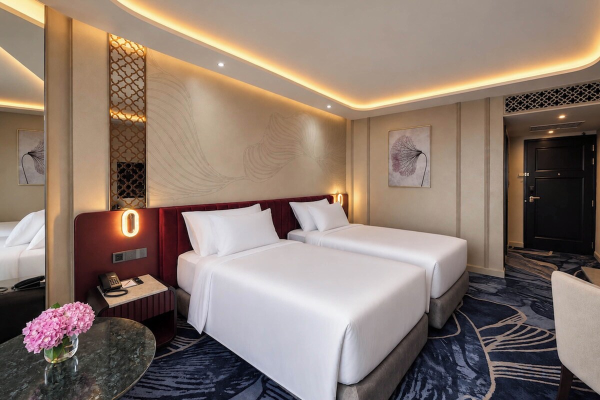 Ein Hotelzimmer mit zwei Betten, einem Schreibtisch, einem Telefon, einer Vase mit Blumen und Bildern an den Wänden.