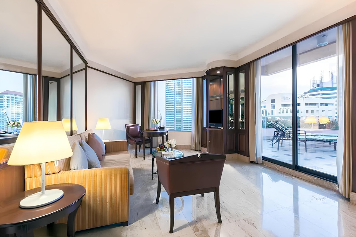 Ein modernes Hotelzimmer mit einer Couch, einem Sessel und einem kleinen Tisch mit Lampe. Ein großes Fenster bietet Blick auf die Stadtlandschaft.