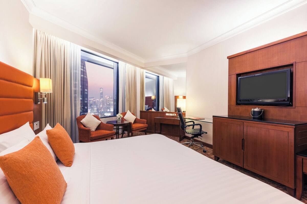 A hotel room with a large bed, a desk, a TV, and a view of the cityscape through the windows.