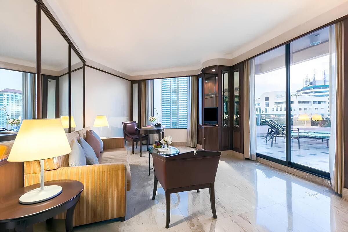 A modern hotel room with a sofa, armchair, and a small table with a lamp. There is a large window offering a view of the cityscape.