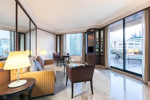 Une chambre d’hôtel moderne, dotée d’un canapé, d’un fauteuil et d’une petite table avec une lampe. Une grande fenêtre offre une vue sur le paysage urbain.