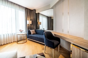 A modern hotel room with a sofa, a side table with a lamp, a wooden desk, and a large window with curtains.