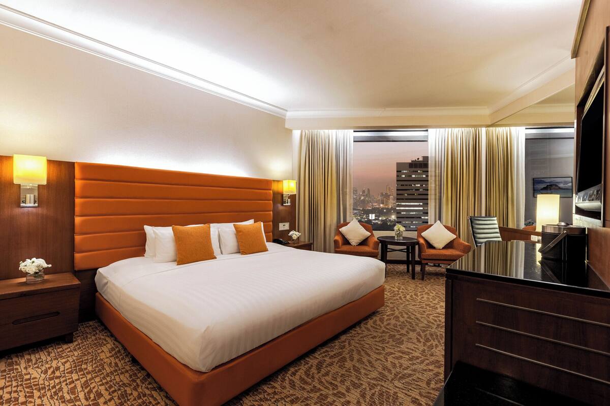 A hotel room with a large bed, a desk, two chairs, a TV, and a city view.