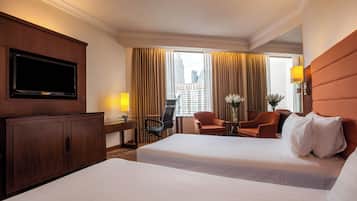A hotel room with two beds, a TV, a desk with a lamp, a chair, and a view of the city.