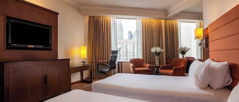 A hotel room with two beds, a TV, a desk with a lamp, a chair, and a view of the city.