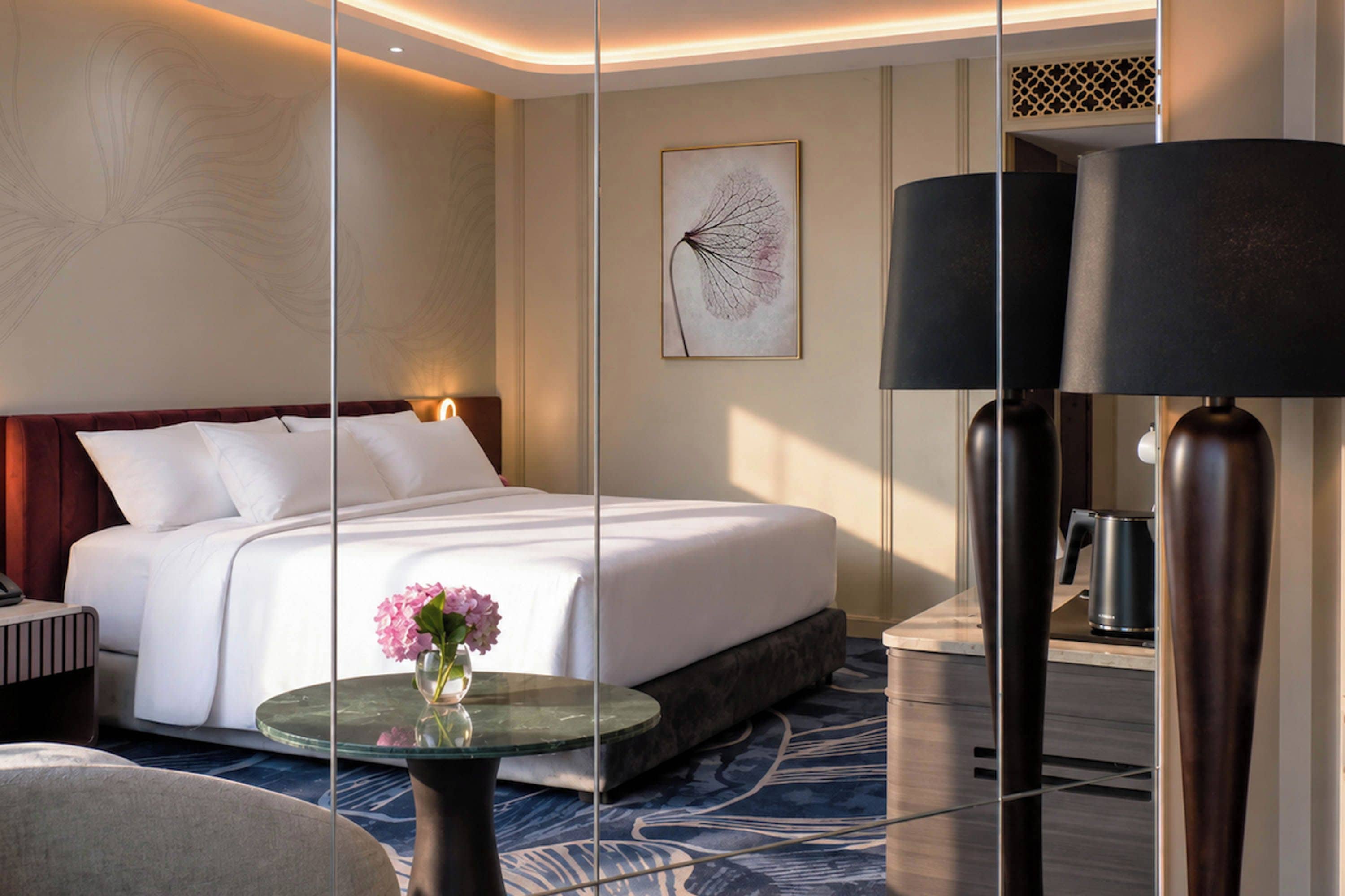 A hotel room with a large bed, a glass coffee table with flowers, a modern lamp, and a framed artwork on the wall.