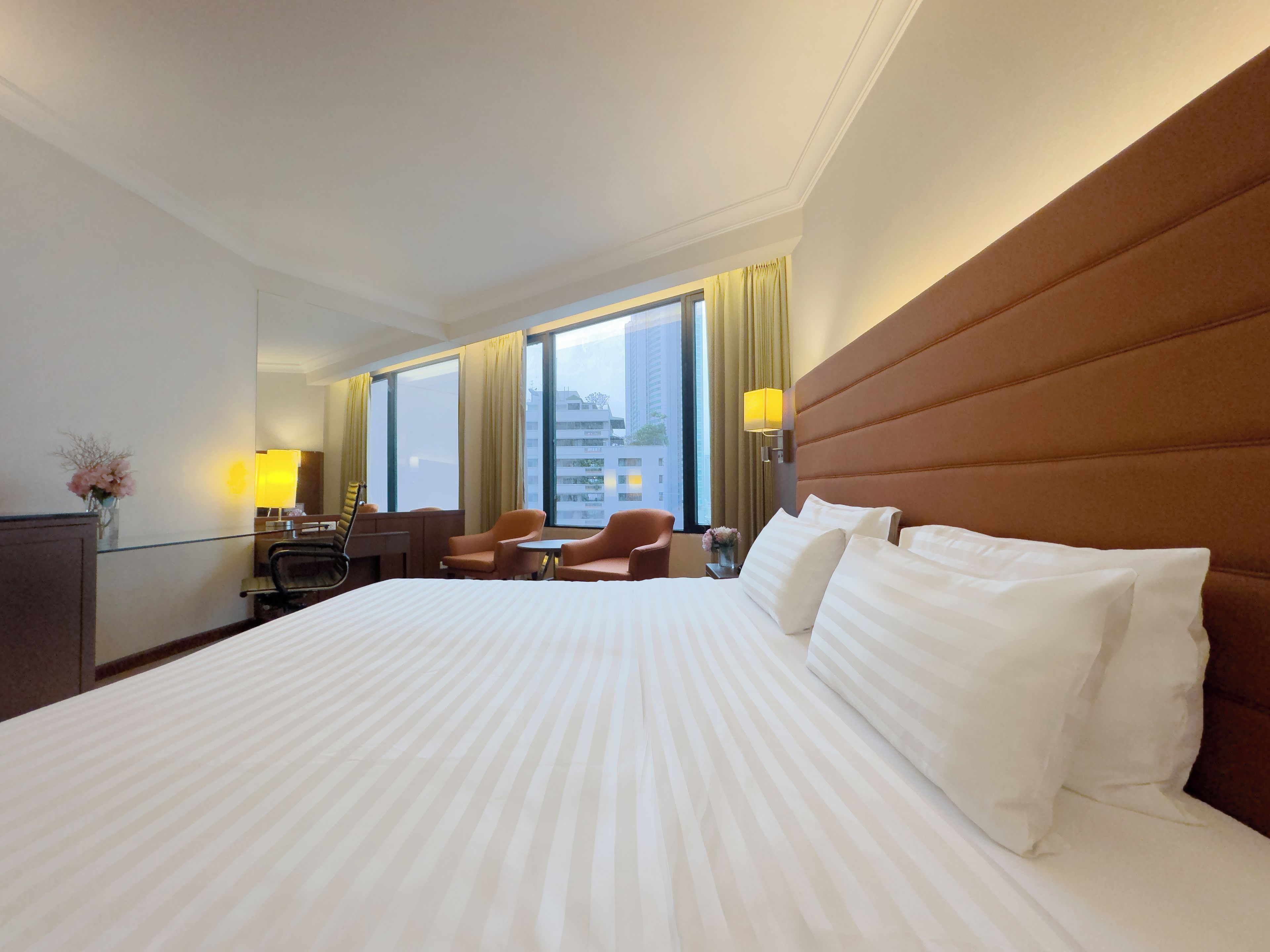 A hotel room with a large bed, a desk, two chairs, a lamp, and a view of buildings through large windows.