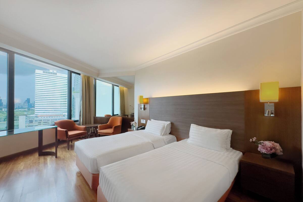 A hotel room with two beds, a desk, and a chair. There is a large window with curtains, a nightstand with a lamp, and a view of the city.