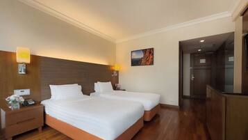 A hotel room with two beds, wooden flooring, a nightstand with a phone, a lamp, and a painting on the wall.