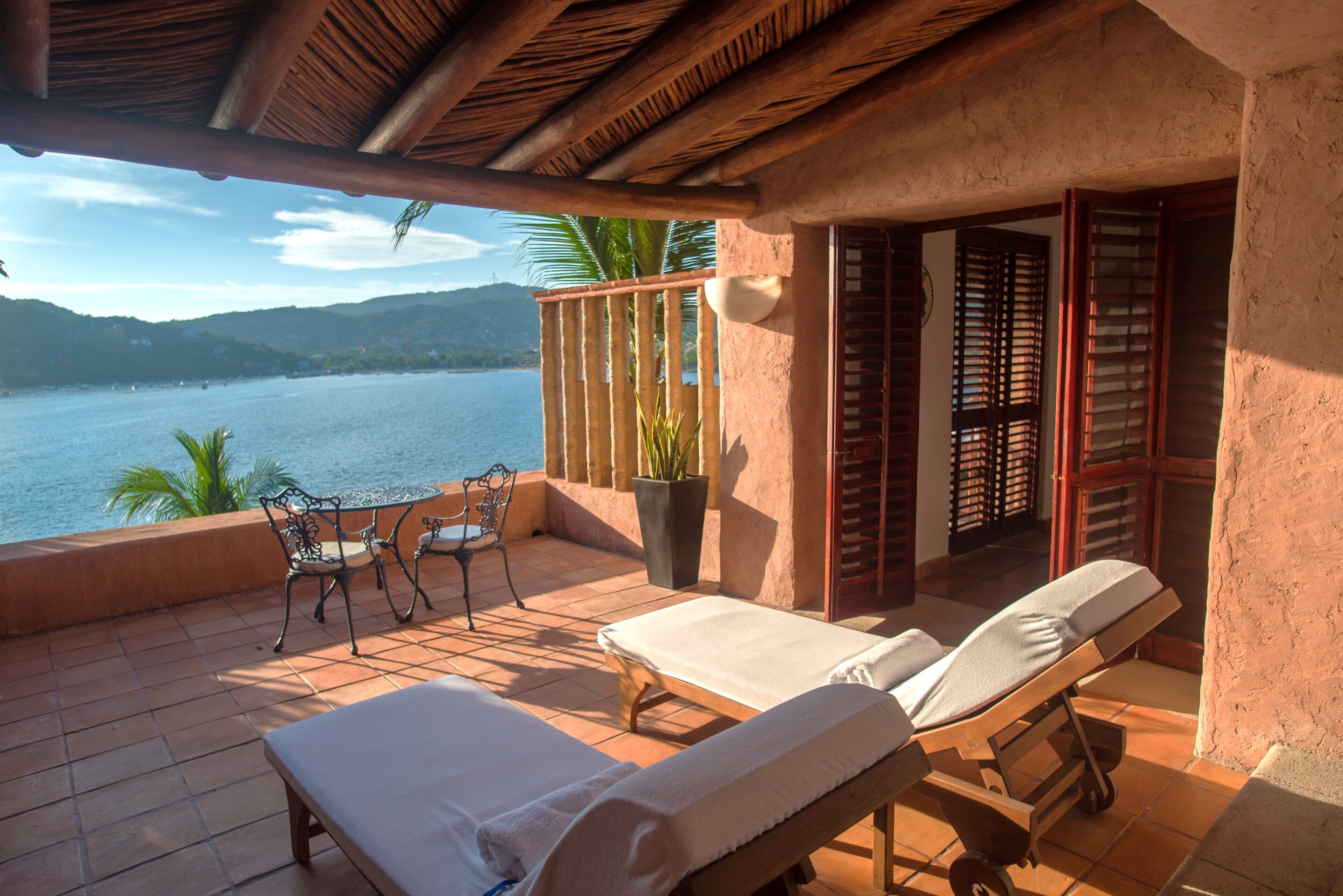 A balcony with two sun loungers, a table, and chairs, overlooking a body of water and hills.