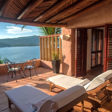 A balcony with two sun loungers, a table, and chairs, overlooking a body of water and hills.