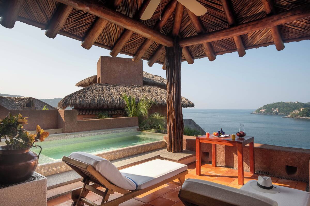 A poolside area with a lounge chair, a table with a vase and fruit, and a view of the sea.