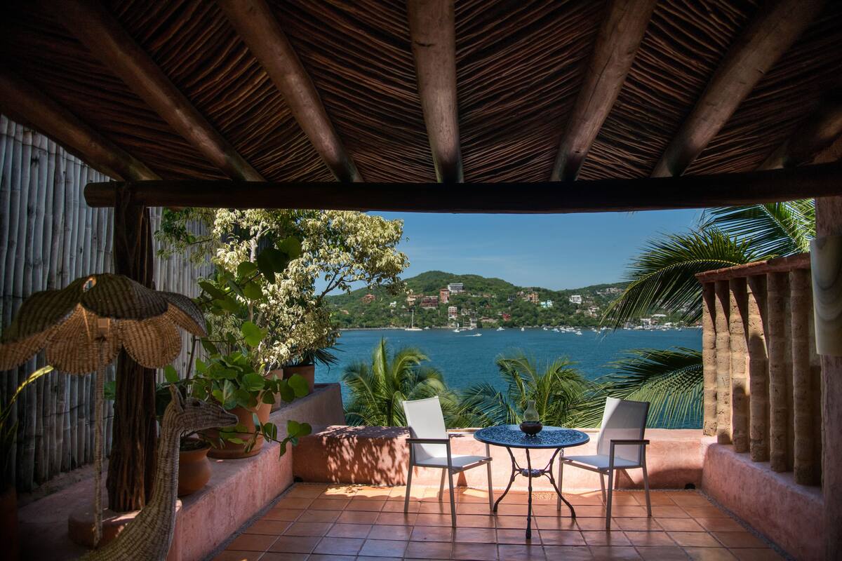 A covered patio with a view of the sea, featuring a table and chairs, potted plants, and a decorative sculpture.