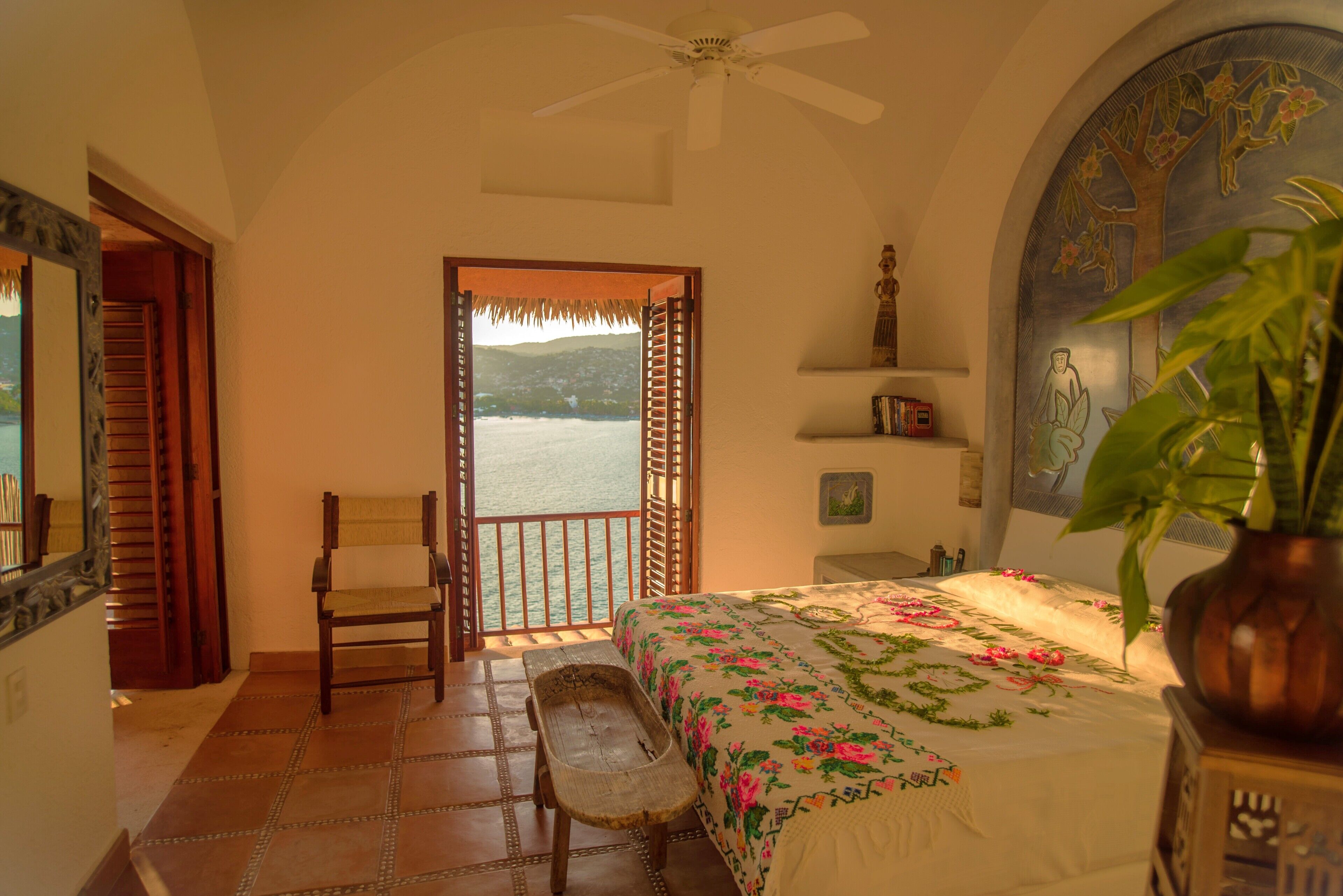 A bedroom with a bed, a chair, a bench, a ceiling fan, a view of the sea, and a decorative wall.