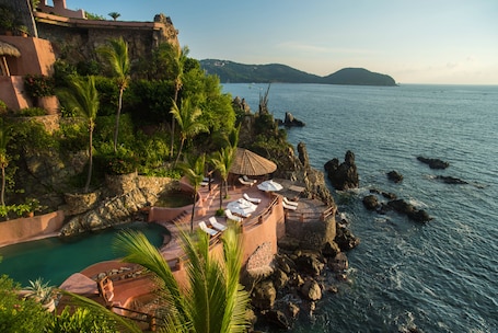 A coastal resort with a pool, lounge chairs, and a view of the sea and distant hills.