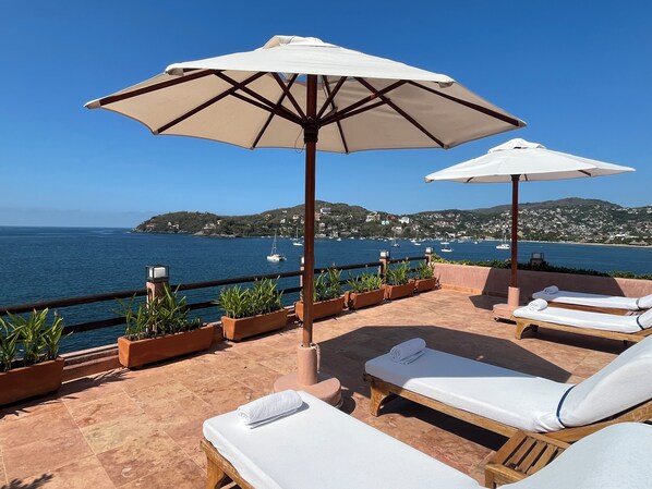 A terrace with lounge chairs and umbrellas overlooking a coastal view with boats and hills. - La Casa Que Canta (Zihuatanejo)